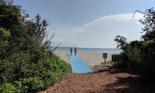 Two people walk along a blue path leading to a sandy beach under a clear sky, surrounded by greenery.