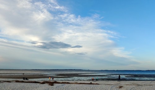 A serene beach scene with a wide sky, low tide revealing sandbars, and a few people exploring the shoreline.