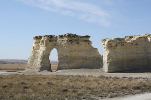 A large rock formation with a natural arch, surrounded by flat grassland under a clear blue sky.