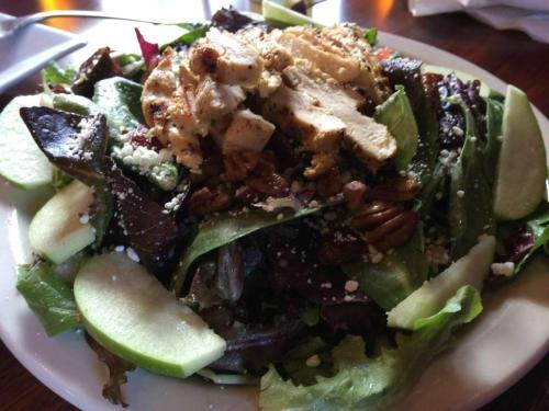 A fresh salad topped with grilled chicken, pecans, feta cheese, and apple slices on a bed of mixed greens.