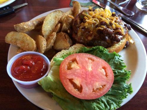 A plate with a cheeseburger topped with chili and cheese, served with potato wedges, lettuce, tomato, and ketchup.
