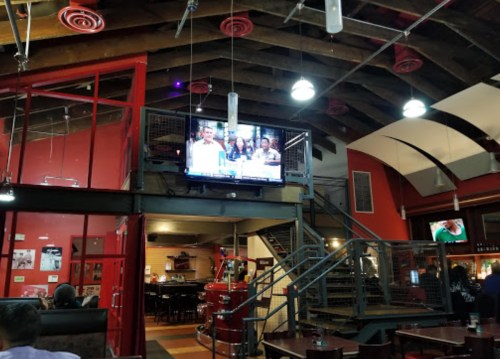 Interior of a restaurant with a TV showing a group of people, featuring exposed beams and a staircase in the background.