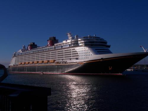 A large cruise ship docked in calm waters under a clear blue sky, reflecting sunlight on the surface.