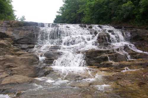 A cascading waterfall flows over rocky terrain, surrounded by lush green trees.