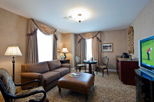 Cozy hotel room featuring a sofa, armchair, table, and TV, with elegant decor and natural light from large windows.