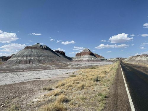 A scenic view of colorful, layered hills beside a deserted road under a clear blue sky with scattered clouds.