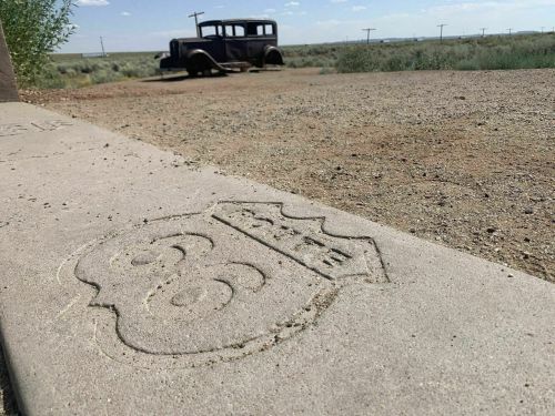 A weathered Route 66 sign embedded in concrete, with an old car in the background and a desert landscape.