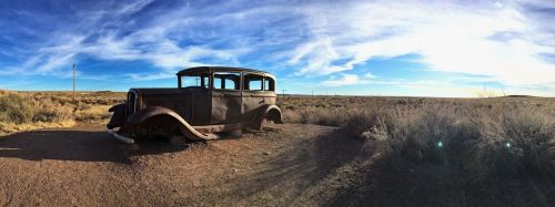 A vintage, rusted car sits in a barren landscape under a blue sky with wispy clouds.