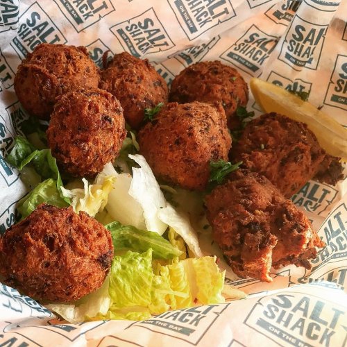 A basket of golden-brown fritters on a bed of lettuce, served with a lemon wedge, all wrapped in branded paper.