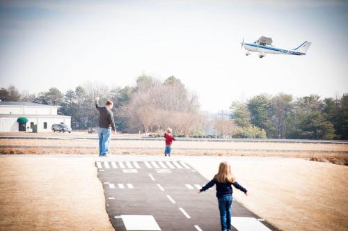A man and two children wave as a small plane flies overhead near a runway on a clear day.