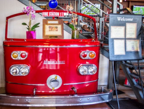 A vintage red fire truck front serves as decor at a restaurant, with flowers and a welcome sign nearby.
