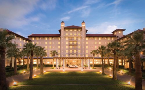 A grand hotel with illuminated windows, surrounded by palm trees and a landscaped entrance under a twilight sky.