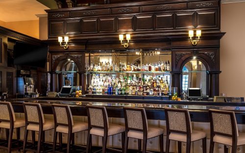A wooden bar with high stools, showcasing a mirrored backlit shelf filled with various bottles of liquor.