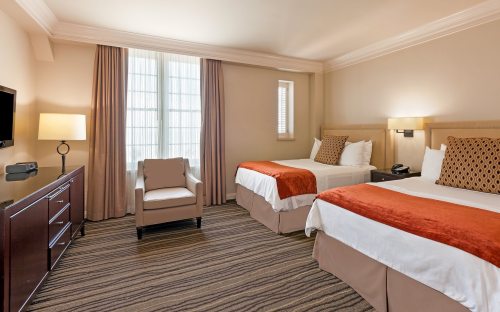 A cozy hotel room featuring two beds, a chair, a TV, and warm-toned decor with natural light from a window.