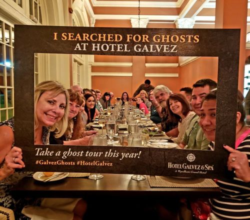 A group of people smiling at a dinner table, holding a frame that says "I Searched for Ghosts at Hotel Galvez."