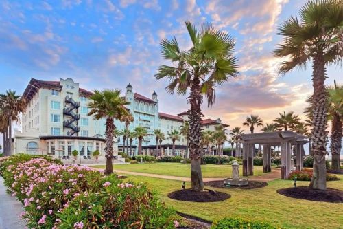 A scenic view of a hotel surrounded by palm trees and colorful flowers under a vibrant sky at sunset.