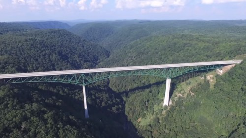 Aerial view of a green bridge spanning a deep valley surrounded by lush green mountains under a blue sky.