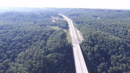 Aerial view of a highway winding through lush green forests and hills under a clear sky.