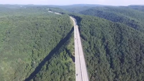 Aerial view of a highway winding through lush green hills and forests under a clear blue sky.