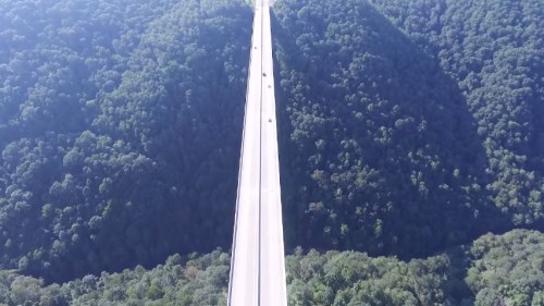 Aerial view of a long, elevated highway surrounded by dense green mountains and trees.