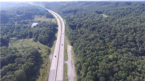 Aerial view of a highway surrounded by lush green trees and a small pond in the distance.