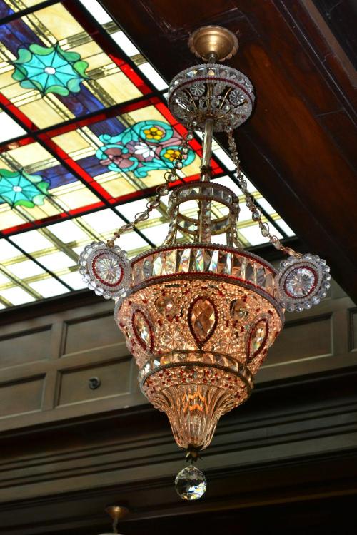 A decorative chandelier with intricate crystal details hangs beneath a colorful stained glass ceiling.