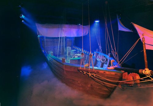 A wooden ship model with sails, surrounded by fog and illuminated by blue lights in a dimly lit space.