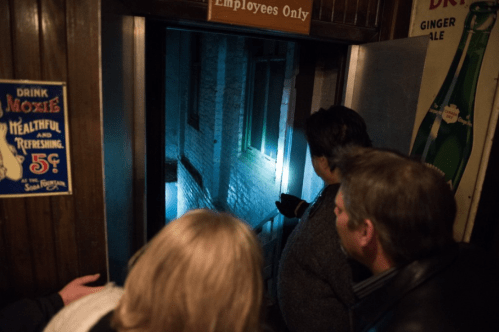 A group of people peering through a doorway into a dimly lit alley, with one person holding a flashlight.