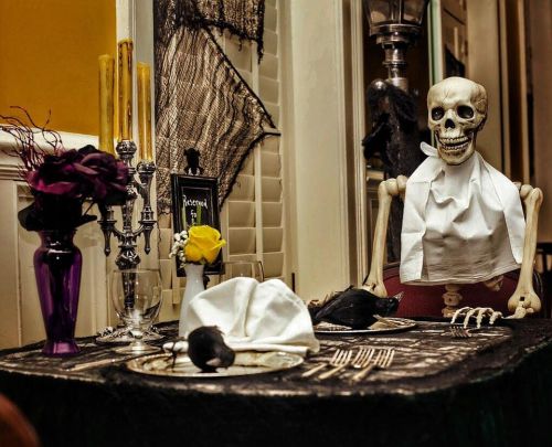 A skeleton sits at a spooky dinner table, adorned with candles, flowers, and a black tablecloth for Halloween.