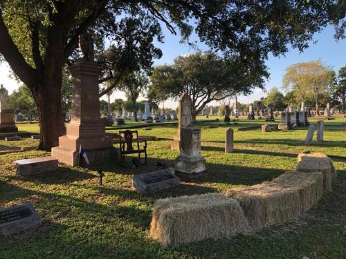A peaceful cemetery scene with gravestones, trees, and hay bales on the grass under a clear blue sky.