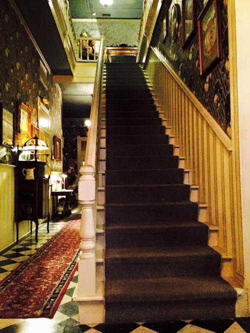 A staircase leads upward in a vintage hallway adorned with framed pictures and patterned wallpaper.