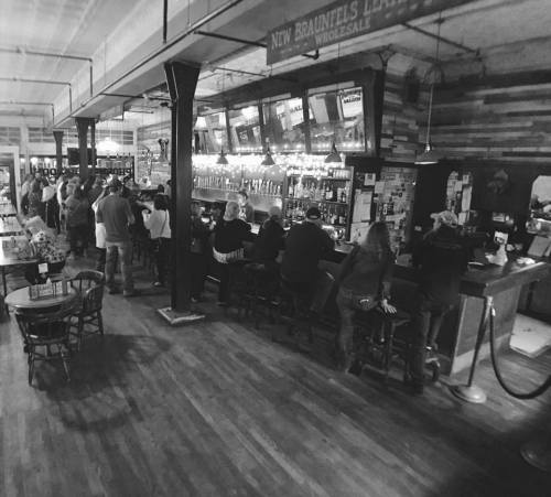 A dimly lit bar scene with patrons seated at the counter and standing, surrounded by wooden decor and shelves of drinks.