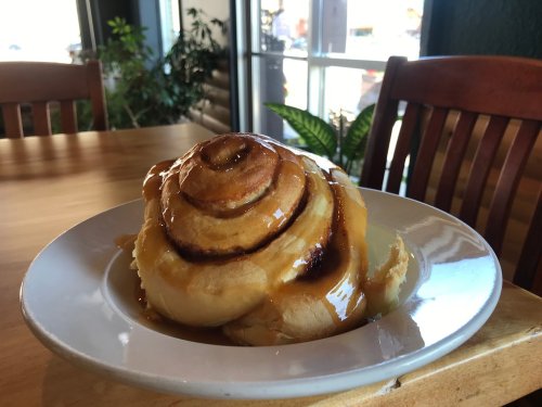 A freshly baked cinnamon roll drizzled with caramel sauce on a white plate, set on a wooden table.