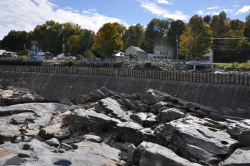 Rocky shoreline with a concrete wall, colorful trees in the background, and houses along the coast under a blue sky.
