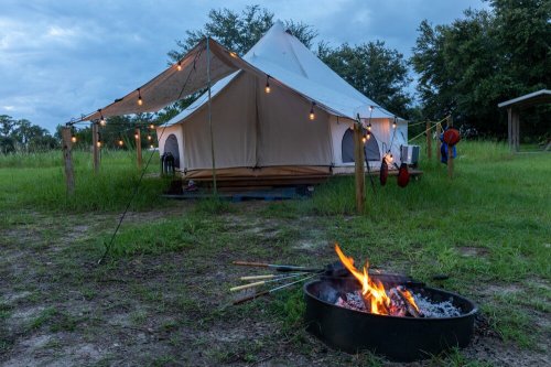 A glamping tent with string lights sits on grass, beside a fire pit with burning logs and skewers in the foreground.