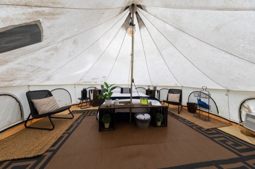 Interior of a spacious tent featuring a table, chairs, plants, and decorative rugs, with soft lighting above.