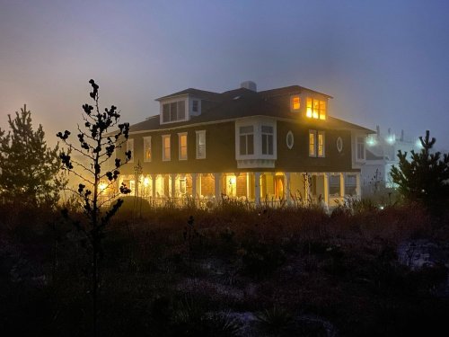 A large house illuminated at night, surrounded by fog and tall grass, with warm lights glowing from the windows.
