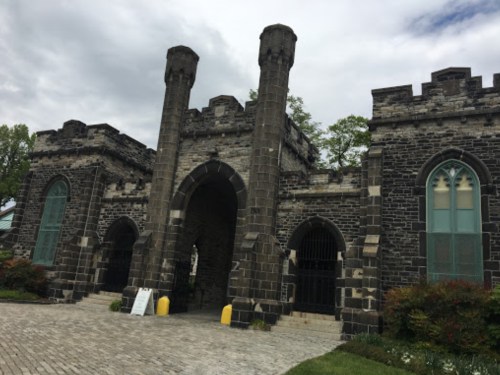 A stone castle entrance with two tall towers and arched doorways, surrounded by greenery and a cobblestone path.