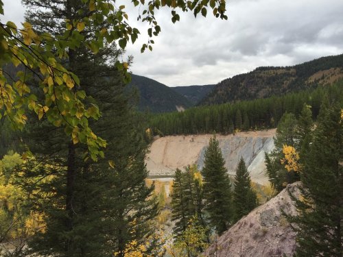 A scenic view of a forested landscape with mountains and a river, featuring autumn foliage and cloudy skies.