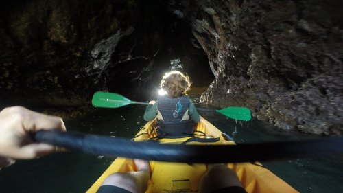 A person kayaking through a dark cave, with sunlight illuminating the entrance ahead. Green paddles are visible.