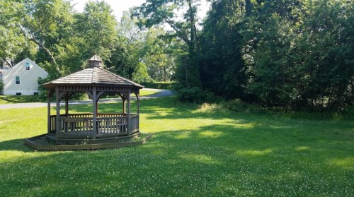 A wooden gazebo sits in a grassy area surrounded by trees, with a road visible in the background.