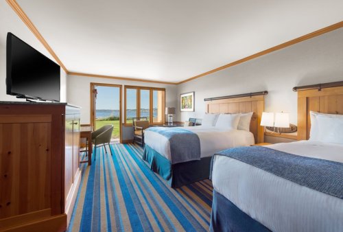 A hotel room with two beds, a TV, a desk, and large windows overlooking a scenic view. Bright and inviting decor.