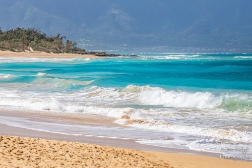 Waves gently crash on a sandy beach, with turquoise waters and lush mountains in the background under a clear sky.