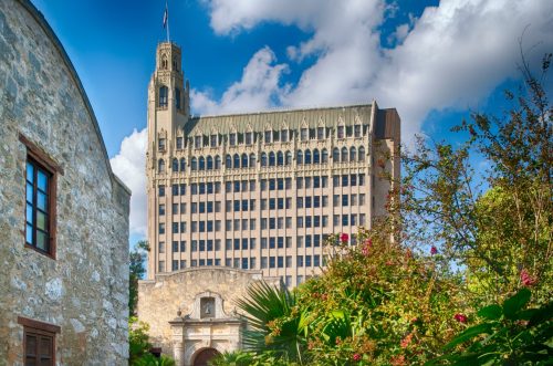 Historic building with a tall tower, surrounded by greenery and flowers, under a blue sky with clouds.