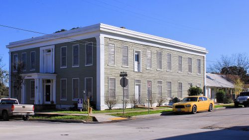 A two-story green building with white trim, surrounded by trees and parked cars on a sunny day.