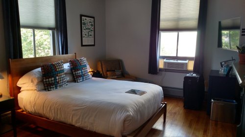 Cozy bedroom with a queen bed, armchair, windows, and a small fridge, featuring warm wood tones and natural light.