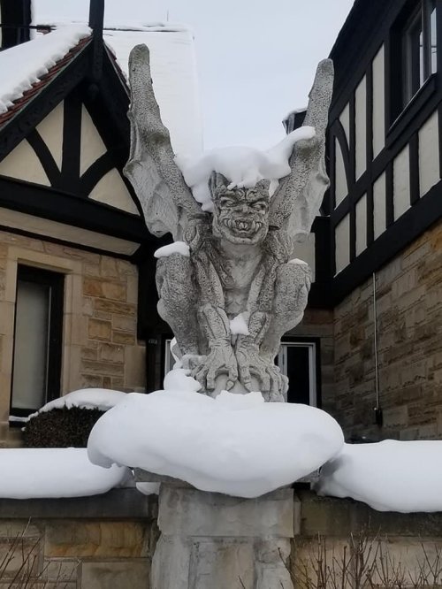 A stone gargoyle covered in snow, perched on a pedestal outside a building with a Tudor-style architecture.
