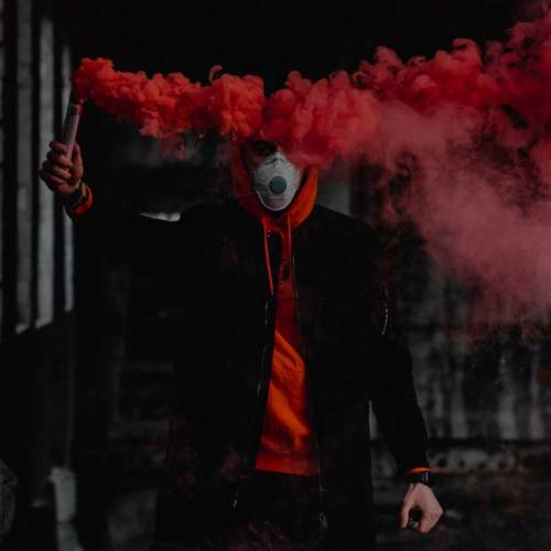 A person in a mask holds a red smoke flare, surrounded by vibrant smoke in a dimly lit industrial setting.
