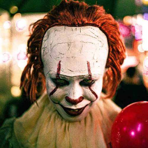 A creepy clown with white face paint, red hair, and a sinister smile, holding a red balloon in a dimly lit setting.