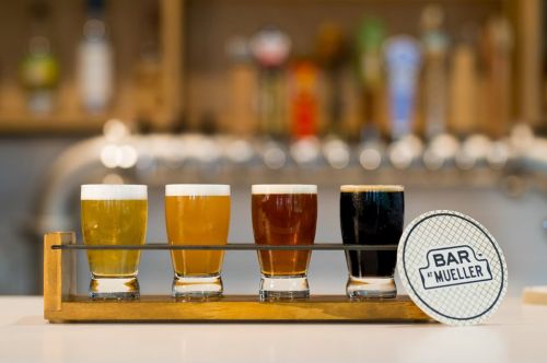 A wooden flight holding four small glasses of different beers, with a coaster labeled "BAR MUELLER" beside it.